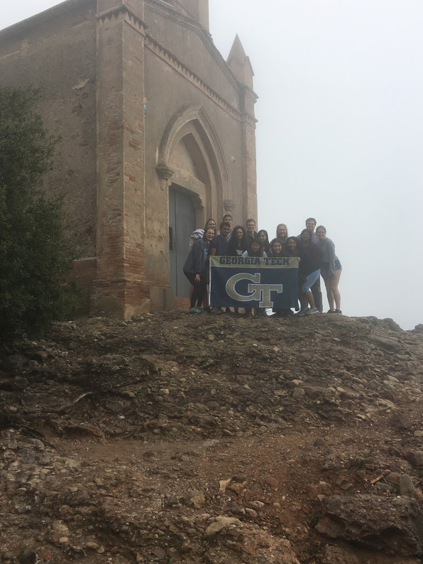 A group of Tech students holding a Georgia Tech flag stand in front of a gothic building in Montserrat on a foggy day