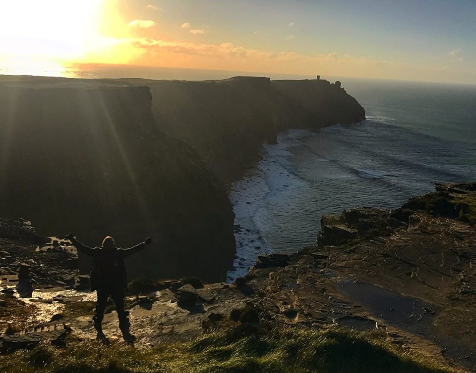 Student standing with arms out-stretched on the Cliffs of Moher with the sun shining over the tops of the cliffs