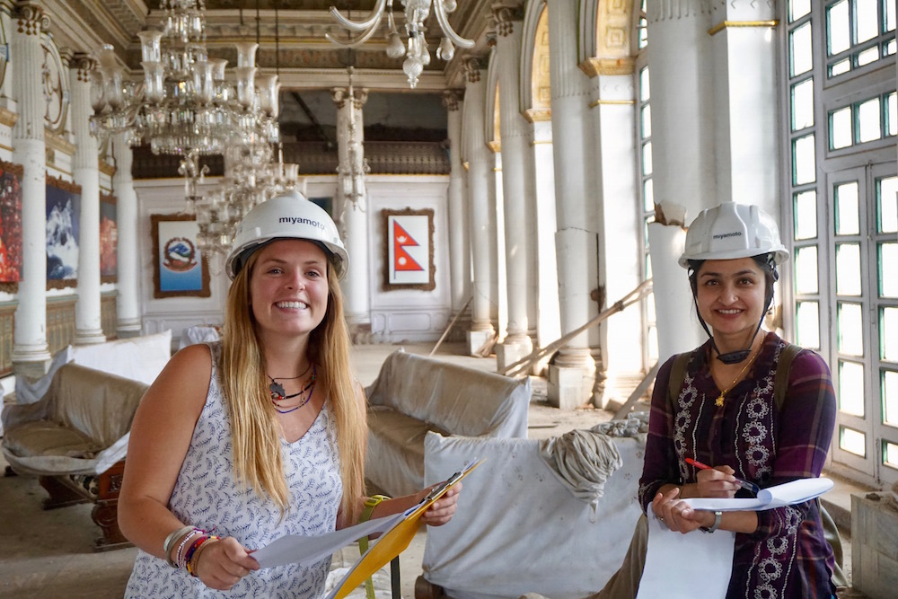Two women wearing hard hats and holding files are standing in an elaborate hall that's being renovated