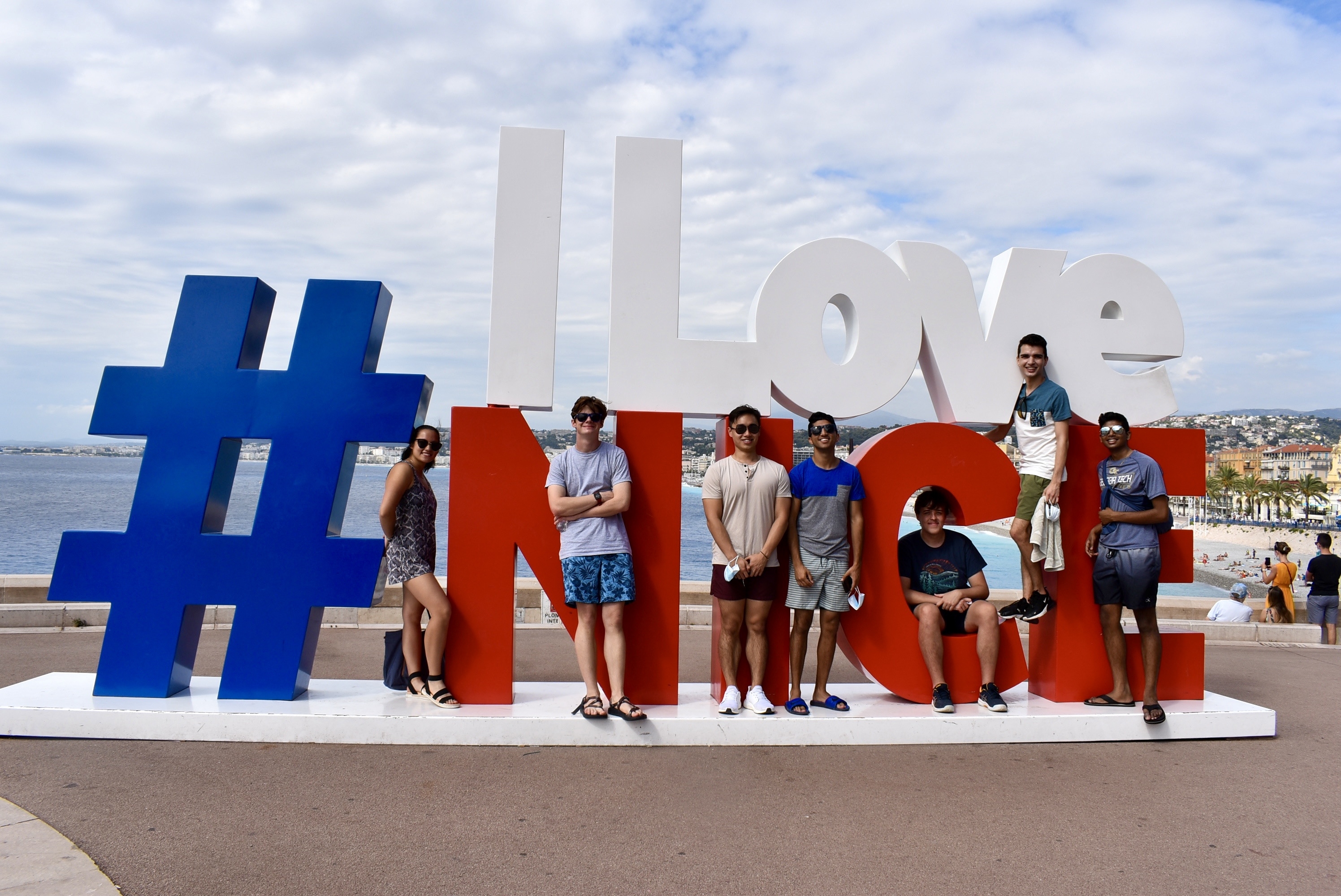 A mixed group of seven GA Tech students stand in a large sign that says, #I Love Nice. The day is sunny with a bright cloudy sky and a body of water in the background