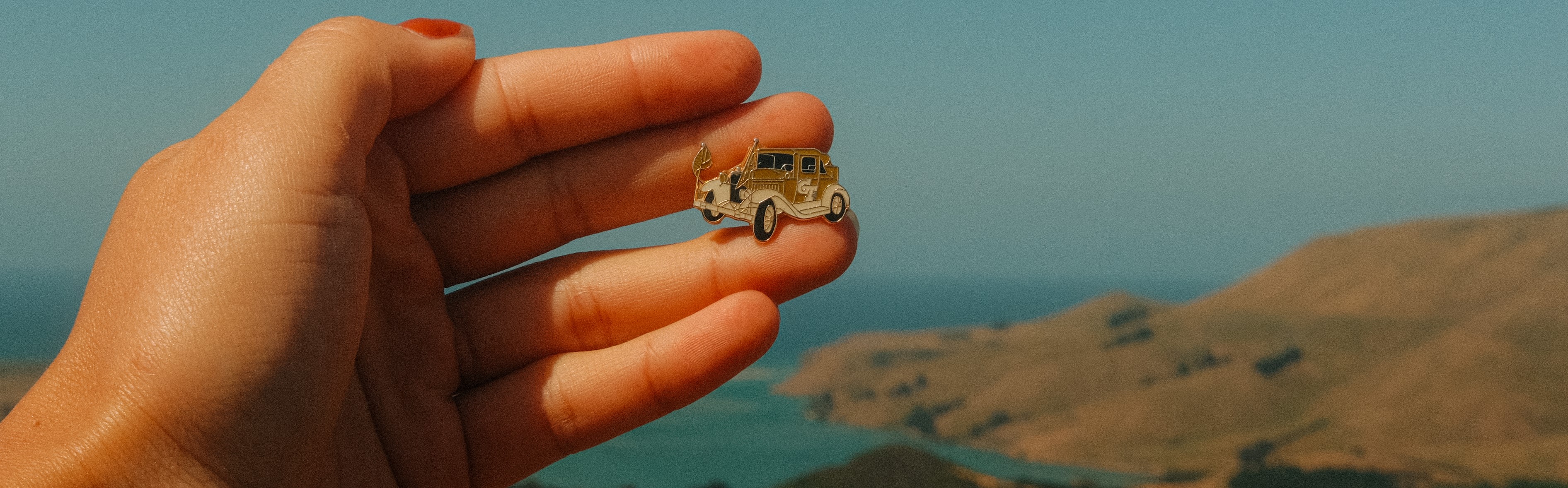 Hand holding a Ramblin Wreck lapel pin in front of a seaside landscape. 